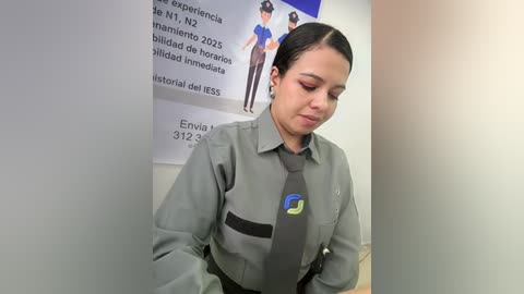 Video of a Latina woman with dark hair, wearing a gray uniform with a blue logo, standing in a white-walled room with informational posters.