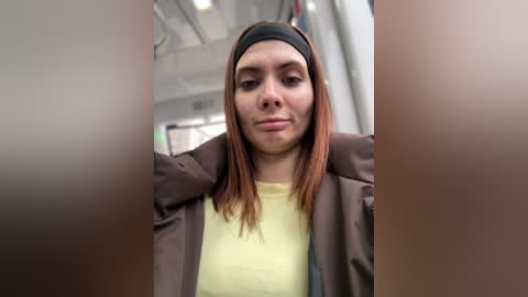 Video of a young woman with straight, reddish-brown hair, wearing a black headband, yellow shirt, and brown jacket, standing in a brightly lit, modern indoor setting.