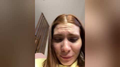 A video shows a young woman with fair skin, straight brown hair, and a yellow sweater, wearing a nose ring. She has a puzzled expression, standing in a dimly lit room with a metal folding chair visible in the background.