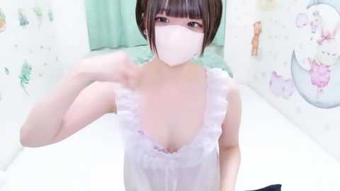 Video of a young Asian woman with short brown hair, wearing a white, frilly camisole, a face mask, and a white sleeveless top, posing in a pastel-themed room with floral and cartoonish wall art.