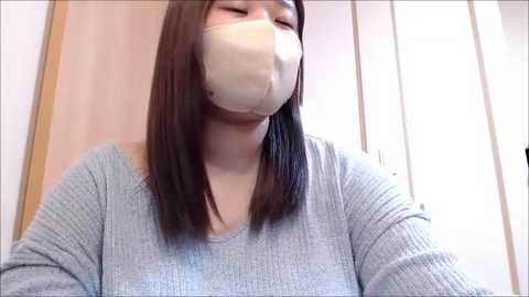 A video of an Asian woman with long, straight, dark hair, wearing a light grey ribbed sweater, a beige face mask, and a white surgical mask. She stands indoors with light wooden and white background.