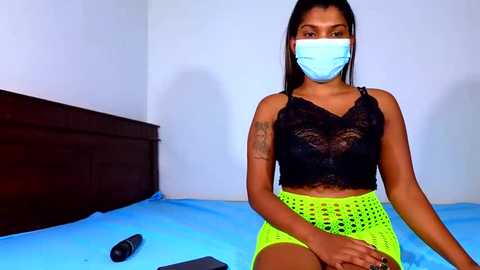 Video of a South Asian woman with medium brown skin, straight black hair, wearing a blue mask, black lace top, neon green shorts, sitting on a blue bed, holding a remote.