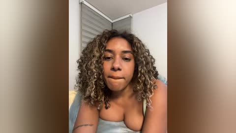 Video of a light-skinned woman with curly, shoulder-length hair, wearing a blue top, sitting indoors. She has a neutral expression. Background includes a white wall with a window and a bed with white sheets.