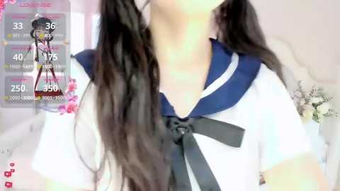 A video of a young woman with long, wavy black hair styled in pigtails, wearing a traditional Japanese schoolgirl sailor uniform with a black bow. The background features a soft-focus, bright room with a white headboard and floral decorations.