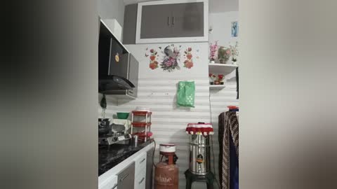 A video of a small, modern kitchen with white wall panels, black appliances, and floral wallpaper. The space is cluttered with utensils, a microwave, and a hanging towel.