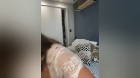 Video of a woman lying on a bed, covered in a white plastic sheet, with a grey and white patterned pillow and grey wall in the background.