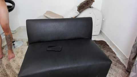 A video of a black leather recliner with a remote control placed on the armrest, surrounded by a white wall, a brown and white rug, and a beige cushion on the floor.