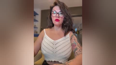 Video of a curvy woman with dark hair and glasses, wearing a white crochet top, revealing tattoos on her right arm, standing indoors with modern furniture in the background.