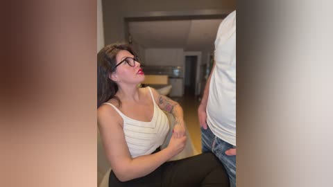 A video of a fair-skinned woman with long brown hair, wearing glasses and a white tank top, kneeling in a modern hallway, looking up at a man in a white shirt.