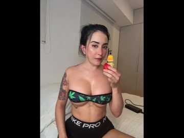 Video of a fair-skinned woman with dark hair, wearing a green camouflage bandeau top and black high-waisted underwear with \"IKE PRO\" branding. She holds a red strawberry in her right hand, sitting on a bed in a modern, minimalist bedroom.