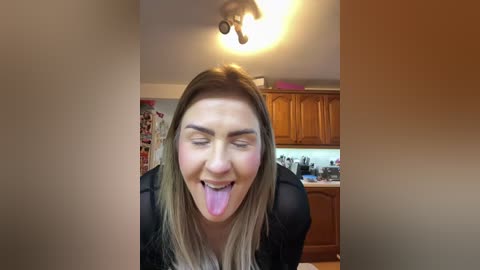 Video of a young woman with blonde hair, wearing a black top, sticking her tongue out playfully in a kitchen with wooden cabinets and a hanging light fixture.