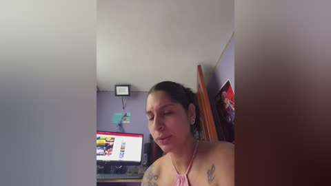 Video of a Latina woman with medium skin tone and dark hair, wearing a pink halter top, sitting in a dimly lit room with a TV displaying a colorful screen, framed by a wooden door and purple wall.