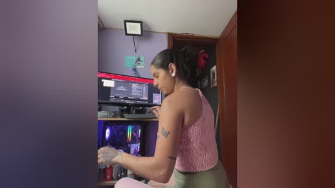Video of a woman with dark hair in a ponytail, wearing a pink sleeveless top and green pants, playing a video game in a cluttered room with a TV and gaming setup.