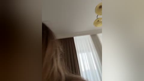 A video shows a blurred figure in a dimly lit room with beige walls, a white curtain, and a yellow light fixture.