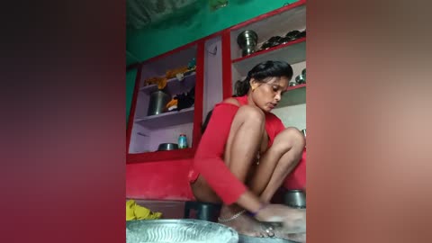 Video of a young woman with medium brown skin, sitting on the floor in a small, brightly colored kitchen. She wears a red top and shorts, and is focused on her task.