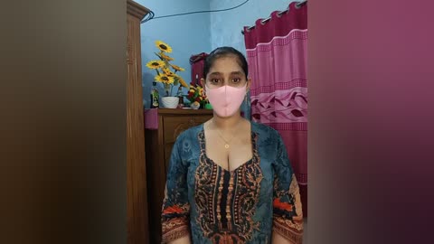 A video of a woman wearing a pink mask, sitting indoors. She has a medium build, dark hair, and wears a traditional, patterned blouse. The background features a wooden table with flowers, pink curtains, and a blue wall.