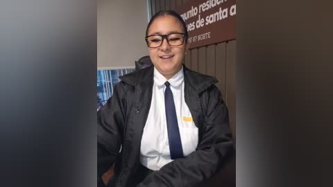 Video of a smiling, middle-aged man with glasses, dark hair, wearing a black jacket, white shirt, and blue tie, sitting in a restaurant with a Spanish sign behind him.
