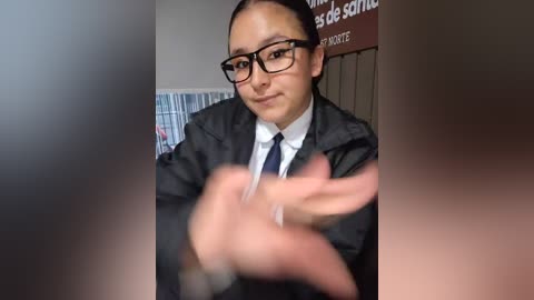 Video of a young, dark-haired woman in glasses and a dark suit, gesturing with her hands, set against a blurred background with a partial text sign.