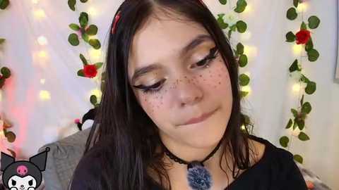 Video of a young woman with fair skin, long dark hair, and pronounced freckles. She wears a black top and a choker necklace. Background features fairy lights and green leaves.