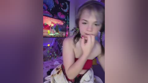 Video of a young, fair-skinned girl with blonde pigtails in a red bikini, biting her finger, in a dimly lit room with a video game TV screen in the background.