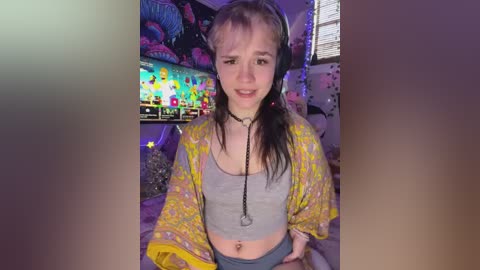 Video of a young woman with fair skin and long brown hair in a ponytail, wearing a yellow floral kimono over a grey crop top and blue shorts, sitting in a colorful, cluttered bedroom with a TV displaying a game.