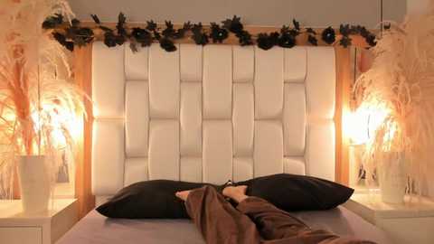 Video of a modern bedroom featuring a tufted white headboard, two black pillows, and a bed with beige and brown bedding. Warm-toned lamps with dried plant accents flank the bed on white nightstands.