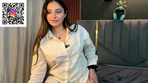 Video of a young woman with long, dark hair, wearing a white button-down shirt, seated on a bed with gray bedding.