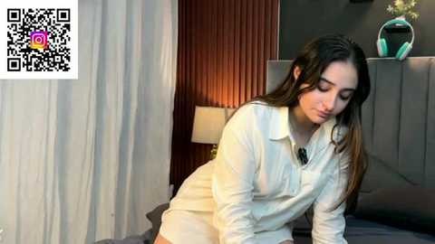 Video of a young woman with long brown hair, wearing a white button-down shirt, leaning forward, in a modern bedroom with a bed, lamp, and curtains. QR code in the corner.