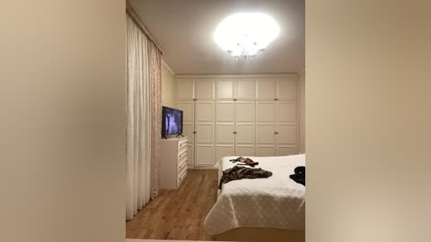 Video of a minimalist bedroom with white walls, wooden floors, and built-in wardrobes. A bed with white linens and a brown blanket is against the right wall. A flat-screen TV and a dresser are on the left.
