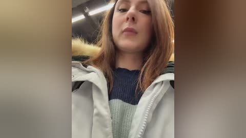 Video of a young woman with straight, light brown hair, wearing a white jacket over a navy-blue sweater. Background shows blurred, indoor environment with a yellow fur trim.