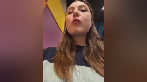 Video of a young woman with fair skin, long brown hair, and closed eyes, wearing a dark blue and light gray striped sweater. Background features a yellow and purple wall.