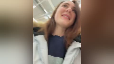 Video of a smiling young woman with shoulder-length brown hair, wearing a blue top under a grey cardigan, in a brightly lit room with white ceiling tiles.