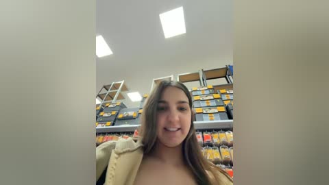 Video of a young woman with long brown hair, smiling, standing in a brightly lit supermarket aisle with fluorescent lights. She wears a beige jacket. Shelves behind her are filled with various products.