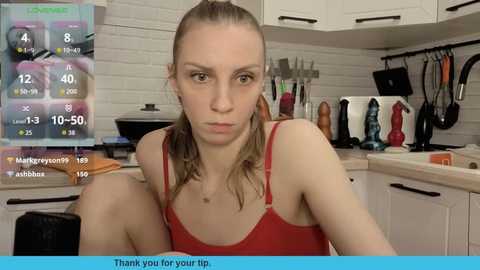 Video of a serious-looking woman with light skin, long blonde hair tied back, wearing a red tank top, sitting in a white kitchen. Background shows utensils and gadgets.