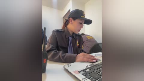 Video of a young woman, possibly Asian, wearing a brown security uniform, cap, and tie, seated at a desk, typing on a laptop, with a blurred background featuring another security guard and office furniture.