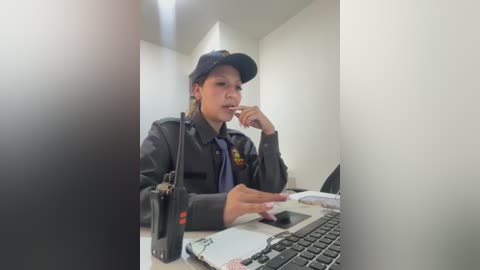 Video of a young Asian woman in a uniform, black hat, and glasses, chewing gum, working on a laptop in a minimalist, white-walled office.