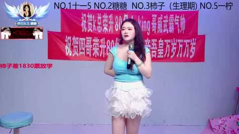 Video of an East Asian woman with long black hair, wearing a turquoise top and ruffled white skirt, speaking into a microphone in front of a red banner with Chinese text, in a brightly lit room.