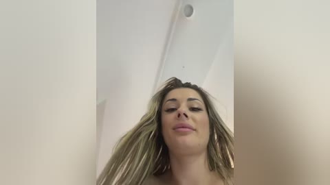 Video of a Caucasian woman with long, straight blonde hair, wearing light makeup, standing in a minimalist room with white walls and a circular light fixture.