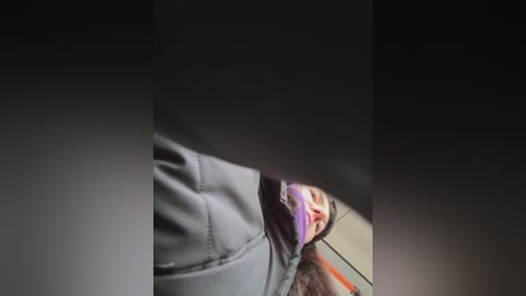 A video showing a woman gagged with a purple ball gag, wearing a grey hoodie, her head tilted back, in a dimly lit, possibly indoor setting.