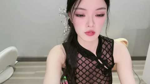 Video of an Asian woman with long black hair, wearing a sheer black fishnet top, sitting in a modern, minimalistic room with white furniture and light wood flooring.