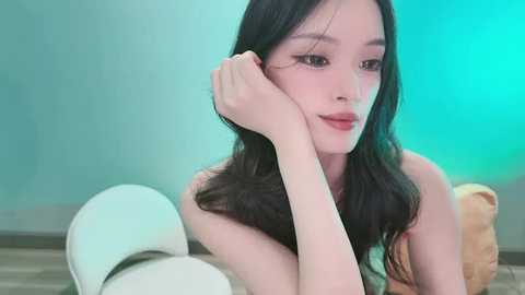 Video of an Asian woman with long black hair, fair skin, and minimal makeup, leaning on a white toilet, against a turquoise and beige background, looking contemplative.