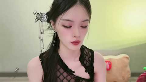 A video of an East Asian woman with pale skin, dark hair styled with a floral hairpin, wearing a black mesh top, in a softly lit room with a stuffed bear.