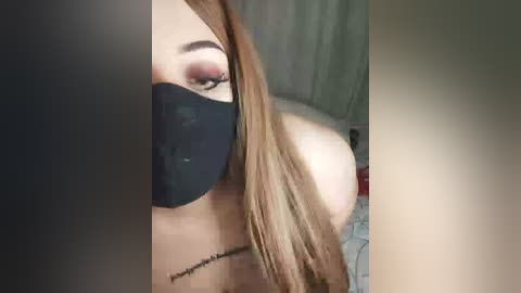 A close-up video of a woman with long, straight brown hair, wearing a black face mask and pink eyeshadow, partially visible in a dimly lit room with greenish curtains.