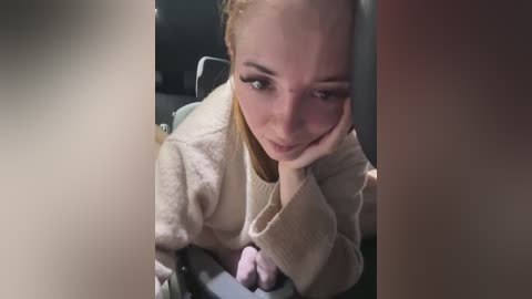 A video of a blonde woman with fair skin, wearing a beige robe, resting her head on her hand, with a soft smile. Background is blurred.