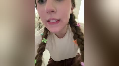 Video of a young Caucasian woman with fair skin, green eyes, and brown hair styled in two braids with colorful beads. She wears a cream-colored shirt and brown skirt, captured from a low angle.