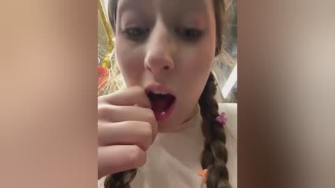 Video of a young woman with fair skin, wearing a white shirt, and brown braided hair. She's licking her finger, with a blurred background featuring yellow and orange flowers.