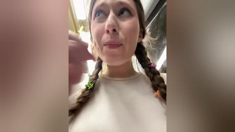 A video of a young woman with fair skin and brown hair styled in two braids, wearing a plain white shirt. She has a slight smile. The background is blurry with a hint of a gray wall.