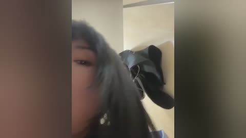 A blurry video shows a partially obscured person with long, dark hair, wearing a black jacket, leaning against a beige wall. A black jacket hangs on a hook behind them.