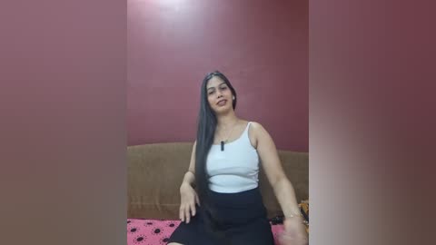 A video of a woman with long, straight, dark hair, wearing a white tank top and black skirt, seated on a pink polka-dotted bedspread against a red wall.