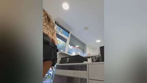 A video showing a person with short, curly brown hair wearing a black T-shirt, standing in a modern, well-lit room with a white desk, monitor, and a black printer.